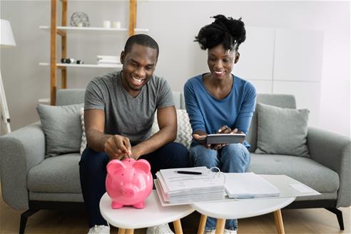 couple sitting on couch putting in money in savings piggy bank on coffee table